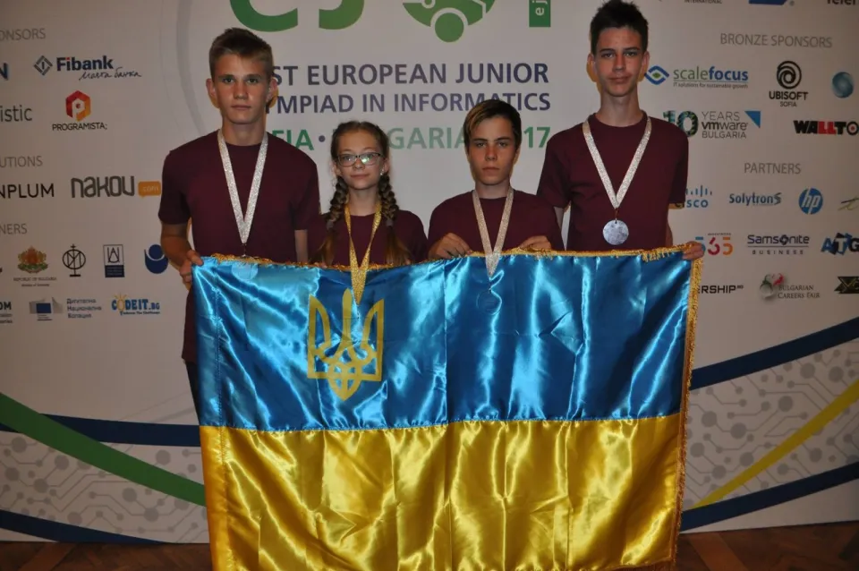 Four members of Ukraine’s team in maroon T-shirts with their medals holding the Ukrainian flag in front of the first EJOI’s banner in Sofia