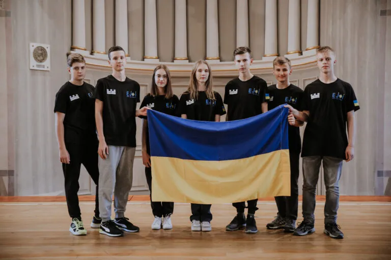Seven members of the team in black EJOI T-shirts holding the Ukrainian flag in a hall with columns