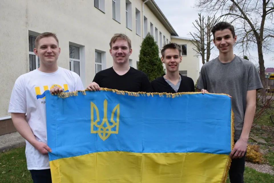 Four members of Ukraine’s team holding the Ukrainian flag in front of a building; one wears a UOI T-shirt