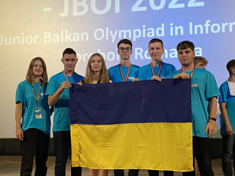 Members of the team with their medals holding the Ukrainian flag on stage; the screen behind reads “JBOI 2022, Junior Balkan Olympiad in Informatics”