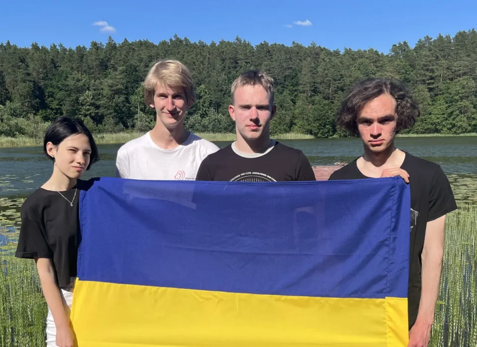 Four contestants holding the Ukrainian flag on a lakeshore in front of a forest